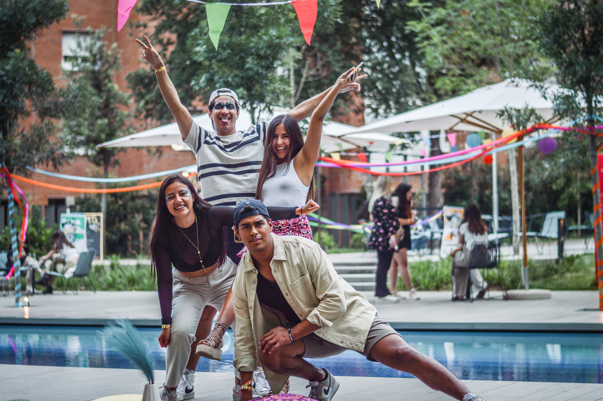 Four people are posed playfully in front of a pool, with colorful flags and decorations in the background. Two are crouching in front, while two stand behind, smiling and making peace signs. Outdoor setting with trees and umbrellas.
